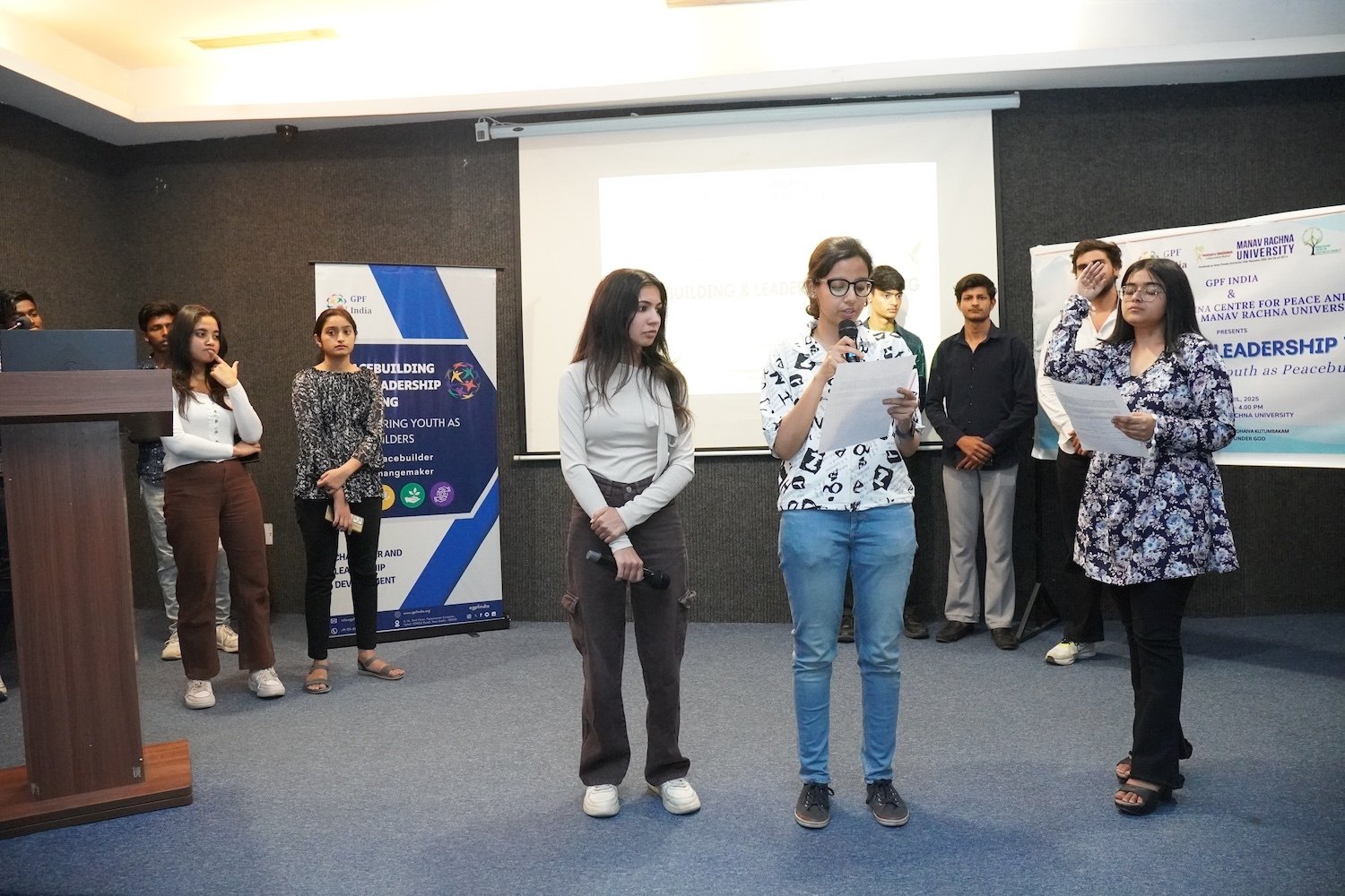 A group of young people stand in a room, embodying peace and leadership as one person speaks into a microphone while others listen. Presentation banners and a projection screen are visible in the background.