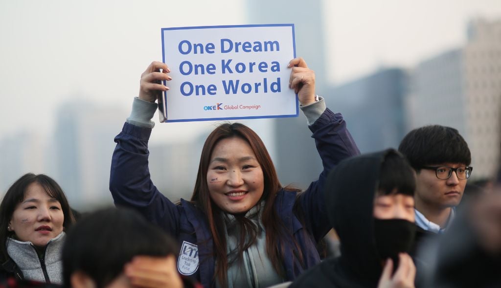 One Korea Membership - GPF (2) A woman in a crowd holds up a sign that reads, “One Dream One Korea One World,” symbolizing the spirit of One Korea Membership. Other people stand around her, some wearing masks and winter clothing.