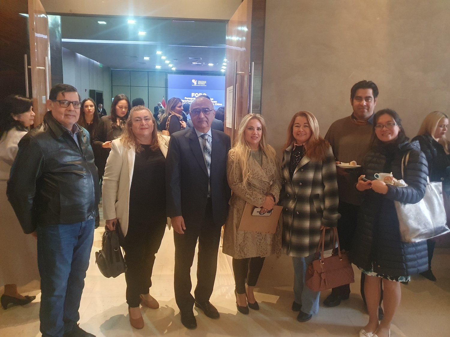 A group of people pose together indoors at a GPF Paraguay event; others are seen in the background with refreshments and a presentation screen, reflecting a commitment to peaceful societies and strong families.