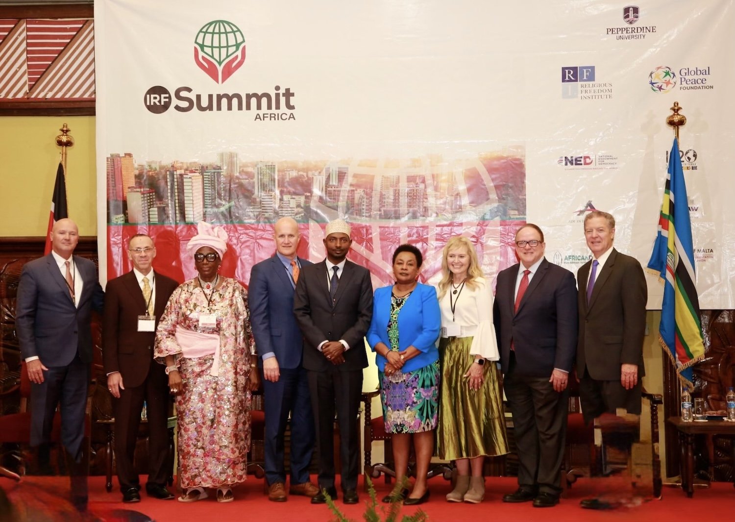 A group of nine people in formal attire stand in front of an IRF Summit Africa banner at an indoor event, celebrating religious freedom and collaboration with the Global Peace Foundation.