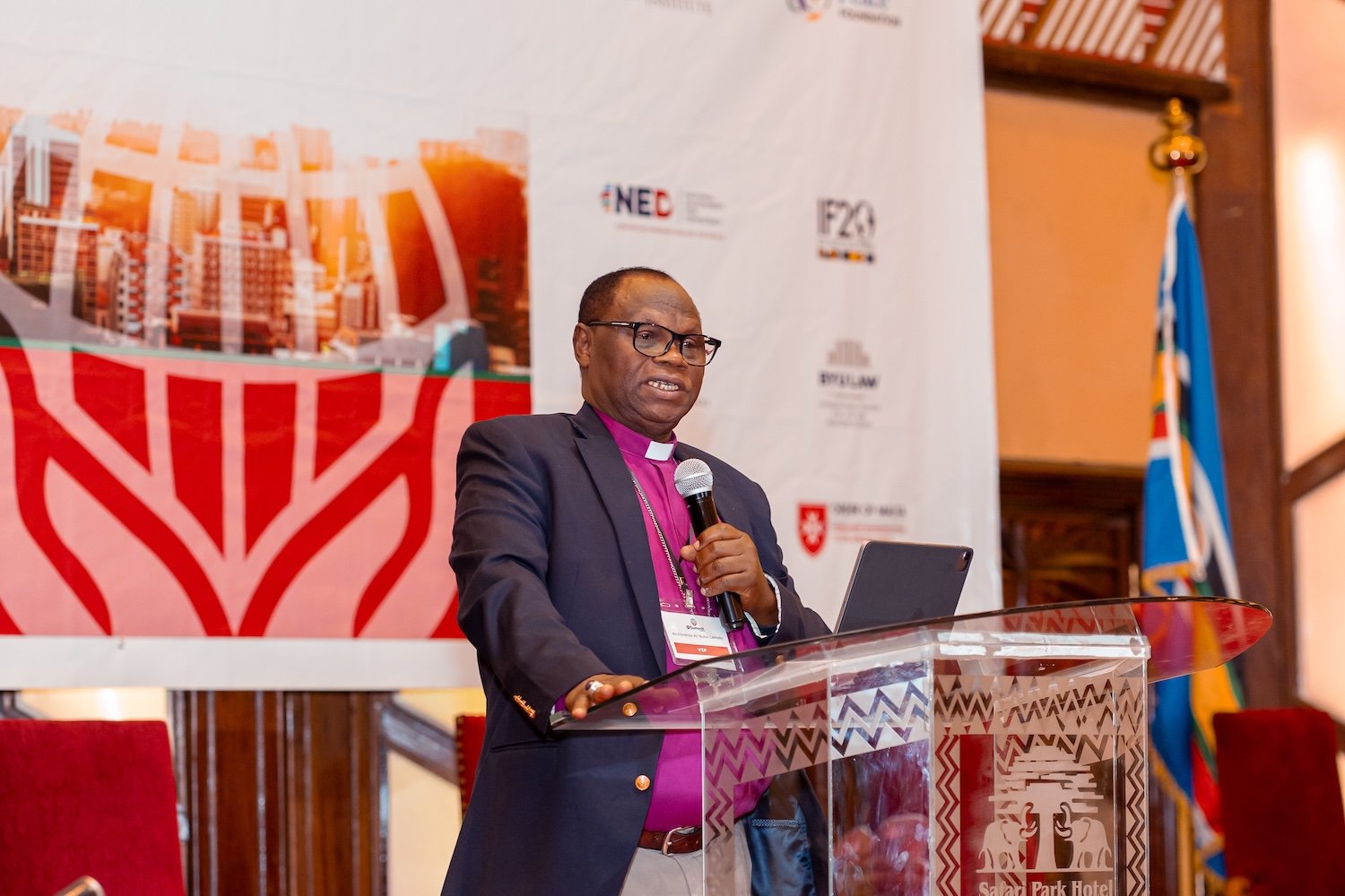 A man in a suit and clerical collar speaks at a podium during the IRF Summit Africa, with a microphone, laptop, and event banner in the background.