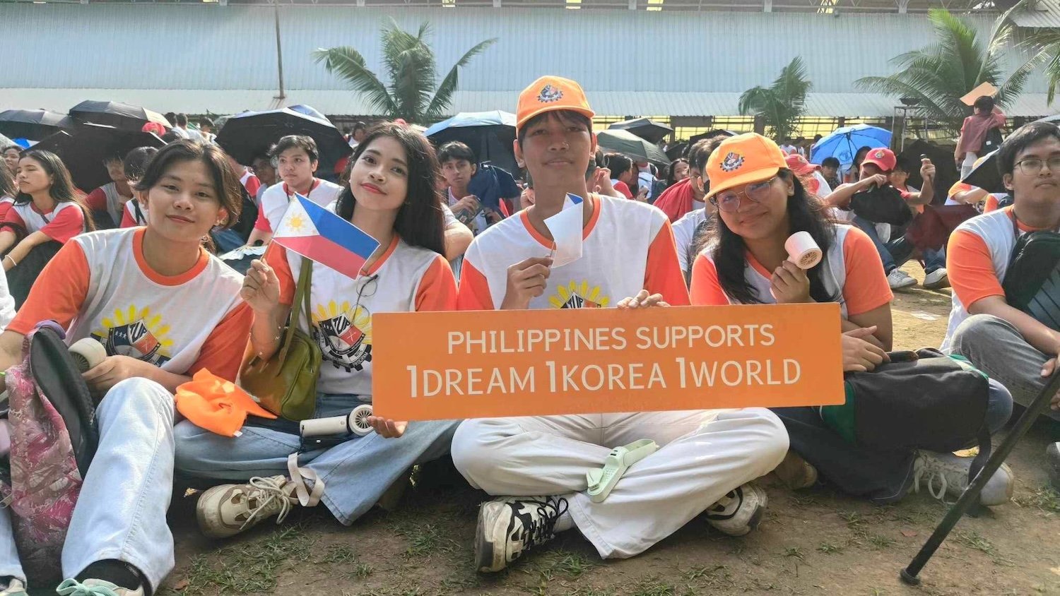 A group of young people wearing matching shirts and caps sit together holding a sign that reads, "PHILIPPINES SUPPORTS 1DREAM 1KOREA 1WORLD," along with small flags, showing their support for Korean Dream and reunification.