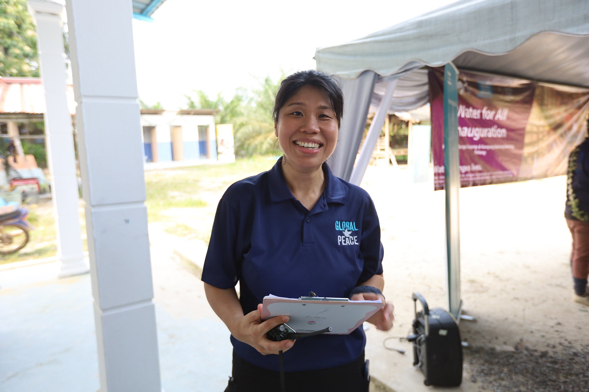 A woman in a navy "Global Peace" shirt representing GPF Malaysia smiles while holding a tablet at an outdoor event with tents and banners, highlighting community support projects.