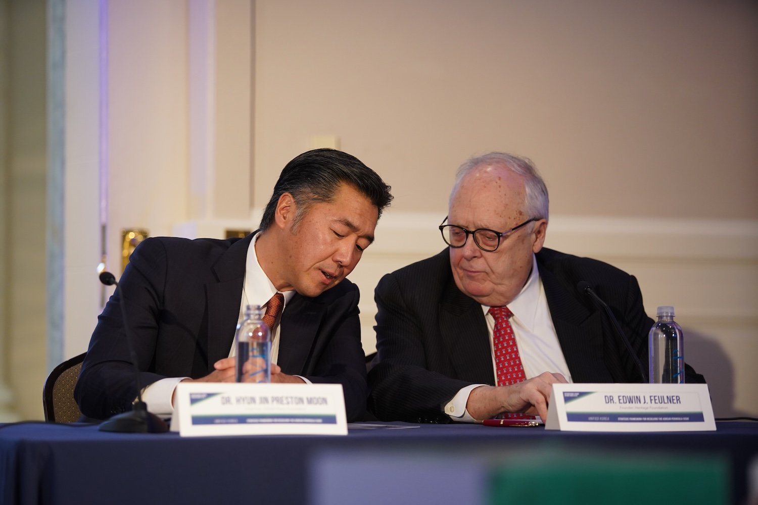 Two men in suits sit at a conference table, leaning in for a private conversation. Nameplates read