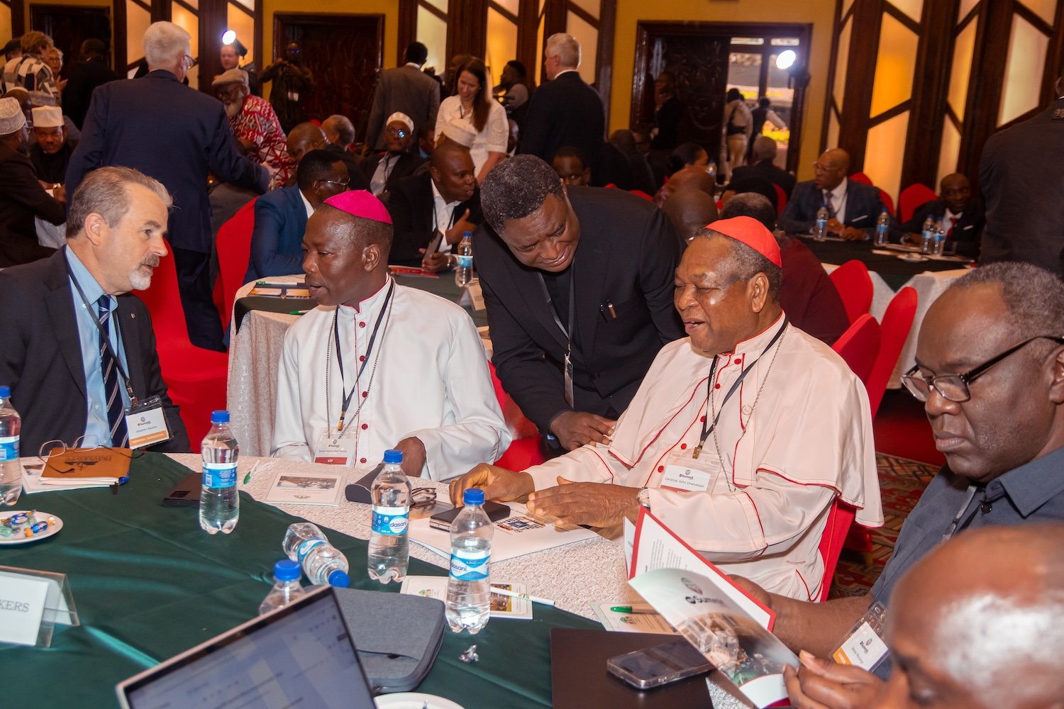 A group of clergy and professionals are seated at a round table, engaged in discussion at the 2025 International Religious Freedom Summit Africa, with documents and water bottles on the table in a large conference room.
